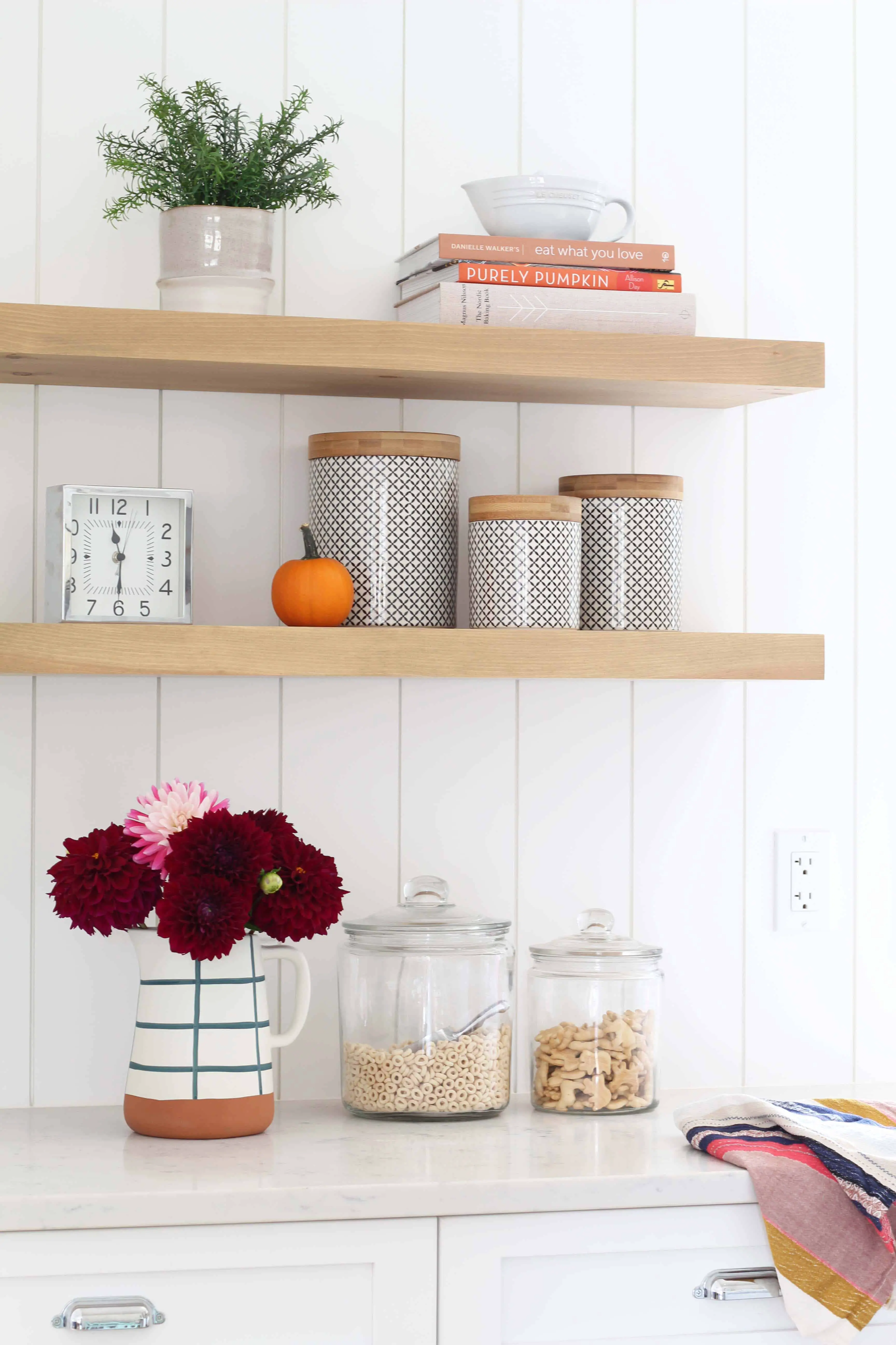 white kitchen with vertical shiplap backsplash, fall mums, purely pumpkin cookbook, wood floating shelves