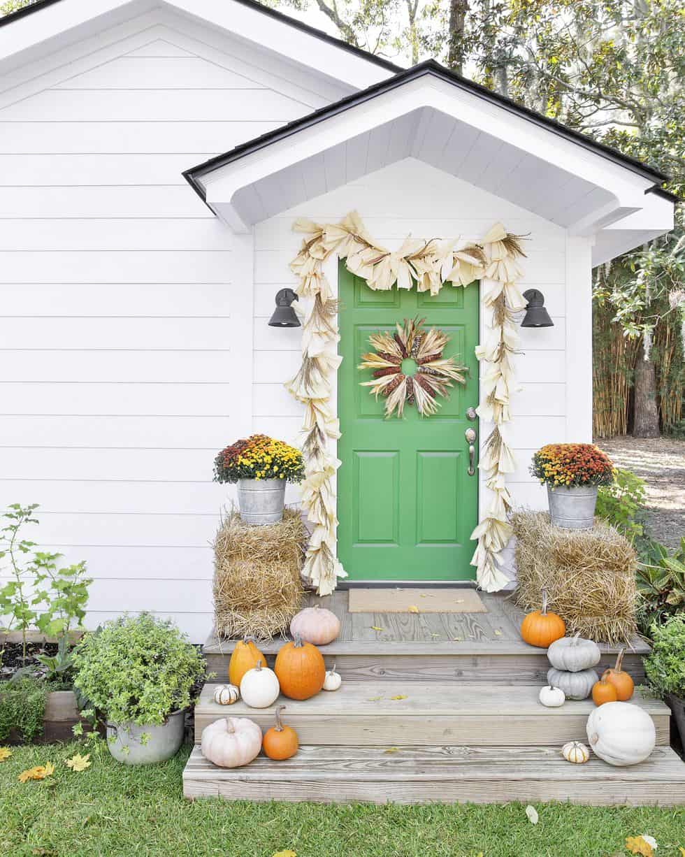 white cottage with green door and pumpkins, mums, and corn husk garland on front porch